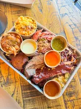 A high-quality, overhead shot of a 'Texas Trinity' BBQ platter featuring brisket, pulled pork, sausage, mac and cheese, and various sides, presented in a rustic tray.