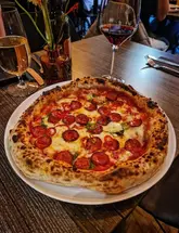 A well-composed, appetizing shot of a Neapolitan-style pepperoni and chilli pizza, centered on a table in a restaurant setting. The image captures a warm, inviting atmosphere with glassware and cutlery, effectively showcasing the food and the dining experience.