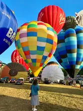 A vibrant, well-composed shot capturing the Bristol Balloon Fiesta, featuring a person standing in the field surrounded by numerous colourful hot air balloons against a clear blue sky.