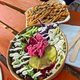 A top-down, outdoor shot of a street food meal featuring a loaded salad bowl with pickles and sauce, accompanied by a side of loaded fries, taken at a food festival.