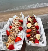 A bright, handheld shot of two loaded waffles on sticks held against a beach backdrop, effectively showcasing the product in a casual, outdoor setting.