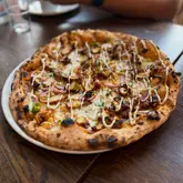 A high-angle, close-up shot of a chicken pizza drizzled with sauce, served on a white plate atop a dark wooden table.
