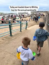 A candid, family-oriented shot capturing children walking down to a beach for a sandcastle competition. The image effectively conveys the atmosphere of a community event, though the background is visually busy.
