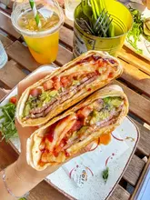 A close-up, first-person perspective shot of a breakfast burrito being held, with a tropical cooler drink and a cafe table setting in the background.