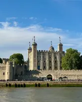 A clear, well-composed photograph of the Tower of London taken from the riverbank. The image captures the historic architecture under a bright blue sky, serving as a straightforward travel or tourism shot.