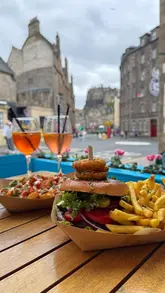 A short, energetic reel featuring a creator enjoying a burger, fries, and cocktails at an outdoor venue in Edinburgh with a view of the castle.