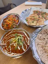 A top-down, flat-lay style photograph of an Indian meal spread, featuring two bowls of curry, a basket of naan bread, and a plate of roti on a wooden table.