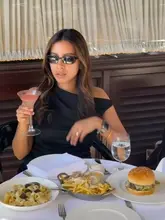 A stylish, well-composed shot of a woman enjoying a meal at a restaurant, featuring a spread of pasta, truffle fries, and a burger, accompanied by a cocktail. The image captures a sophisticated dining atmosphere, suitable for a date night or a social outing.