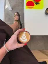 A lifestyle-focused POV shot of a cortado with latte art, held by the creator. The image captures a casual coffee shop moment, emphasizing the drink and the relaxed atmosphere.