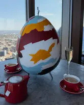 A high-angle shot of an afternoon tea service set against a panoramic city skyline view from a high-rise venue. The table features a large, artistically painted egg-shaped centerpiece, a red teapot, matching teacups, and a glass of champagne, evoking a sophisticated and scenic dining experience.