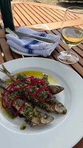 A close-up, appetizing shot of grilled sardines topped with a vibrant green herb salsa and pickled red onions, served on a white plate alongside a glass of white wine on an outdoor wooden table.