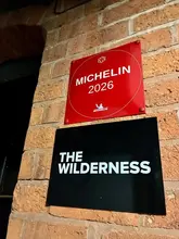 A clear, high-contrast photograph of the exterior signage for The Wilderness restaurant, featuring a Michelin 2026 plaque mounted on a brick wall.