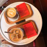 A clean, overhead shot of a flat white and an iced latte served on wooden trays with accompanying tasting notes cards. The lighting is natural and flattering, emphasizing the texture of the coffee and the professional presentation of the venue.