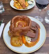 A top-down, eye-level shot of a classic Sunday roast featuring a large Yorkshire pudding, beef, roast potatoes, carrots, and gravy, with a glass of red wine and a partially visible fish and chips dish in the background.