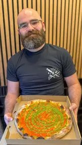 A portrait-style shot of a man holding a takeaway pizza box, showcasing a vodka pesto pizza with a distinct swirl pattern. The setting is a casual restaurant with a simple wooden slat background.