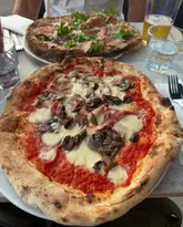 A casual, top-down shot of two Neapolitan-style pizzas on a table. The foreground pizza is the primary focus, displaying toppings such as mushrooms, olives, and ham, while a second pizza and a glass of beer are visible in the background.