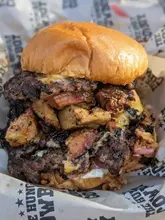 A high-quality, close-up shot of a 'Hungry Cowboy' burger, featuring smashed beef patties, smoked pork, and melted cheese, served on branded paper. The image captures the texture and 'dirty' aesthetic of the street food style perfectly.