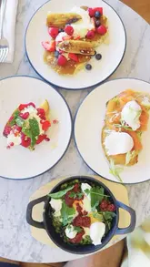 An overhead flat-lay photograph of a brunch spread featuring pancakes, French toast, green shakshuka, and smoked salmon avocado toast on a marble table.