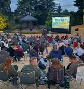 A candid, low-light shot of an outdoor cinema event in a park, showing a crowd of people sitting in camping chairs watching a screen.