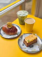 A bright, high-quality product shot featuring a selection of Asian-style bakery items, including a corn creme salt bun, honey toast, and two drinks, set against a vibrant yellow background that aligns with the brand's identity.