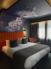 A high-quality, well-composed photograph of a hotel room at Malmaison Deansgate, showcasing the stylish interior design, including the distinctive butterfly-themed ceiling mural and vibrant orange headboard.