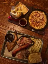 A top-down, slightly noisy shot of a BBQ platter and a pan of mac and cheese on a wooden table, typical of a casual smokehouse setting.