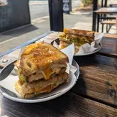 A close-up, appetizing shot of two toasted sandwiches served on parchment-lined plates at an outdoor cafe table. The image highlights the textures of the sourdough, melted cheese, and fresh ingredients, effectively capturing a casual brunch atmosphere.
