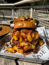A close-up, high-angle shot of a loaded mac and cheese burger served on a branded tray, likely at a casual burger restaurant. The image focuses on the indulgent, messy texture of the food, highlighting the bacon and cheese elements.