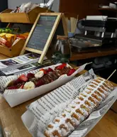 A close-up, eye-level shot of two waffle sticks served on a wooden surface. One waffle is plain with powdered sugar, while the other is topped with Nutella, strawberries, raspberries, and banana slices. The setting appears to be a casual coffee stall or food truck.