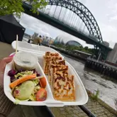A casual, first-person perspective shot of a takeaway container filled with Turkish gozleme, held against the backdrop of the Tyne Bridge at the Newcastle Quayside market.