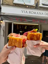 A high-quality, bright POV shot of two people holding up fresh Italian sandwiches in front of the Via Romana Deli storefront in Richmond, effectively showcasing the product and the venue.