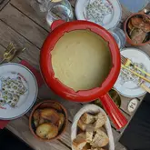 An overhead, flat-lay shot of a cheese fondue set on a wooden table, accompanied by a basket of bread and a bowl of roasted potatoes, captured in an outdoor setting.