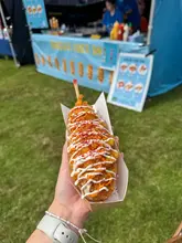 A handheld, close-up shot of a Korean corn dog topped with mayonnaise and chili flakes, captured at an outdoor food festival.