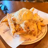 A close-up, high-quality shot of a portion of truffle fries served in a paper-lined basket, topped with a generous dollop of sauce and grated cheese. The image is well-composed with a shallow depth of field, highlighting the texture of the fries and the creamy topping.