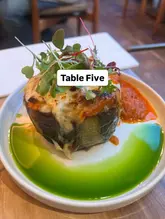 A close-up, top-down shot of a stuffed eggplant dish, likely a moussaka, served on a white plate with a vibrant green oil drizzle and a red sauce, garnished with fresh microgreens.