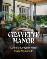 A high-quality, inviting shot of a dining room at Gravetye Manor, showcasing the elegant table settings and the beautiful view of the surrounding gardens through floor-to-ceiling windows. The lighting is warm and natural, creating a sophisticated and tranquil atmosphere suitable for a luxury dining experience.