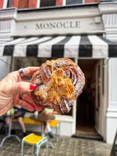 A high-quality, close-up shot of an almond croissant-cinnamon bun hybrid held in front of The Monocle Cafe, with the cafe's exterior and outdoor seating visible in the blurred background.