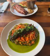 A top-down shot of a dinner table featuring a vibrant risotto dish with green herb oil and a bacon chop in the background. The image captures a casual dining experience at Iris in Wakefield.
