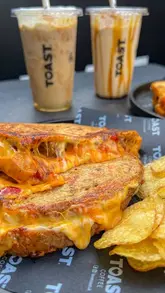 A short, looping video featuring a close-up of a cheese toastie being pulled apart, highlighting the cheese stretch. The creator is visible in the background, and text overlays list 'Basic Human Needs' with the toastie as the third item.