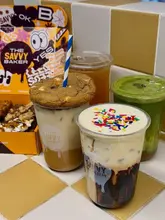 A promotional shot of a summer drink lineup from The Savvy Baker, featuring iced lattes, a matcha drink, iced tea, and a box of brownies.