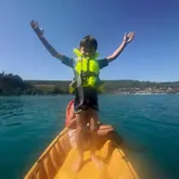 A photo of a young boy standing in a yellow kayak on a lake, arms raised in celebration, capturing a summer holiday activity.