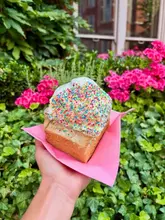 A close-up, handheld shot of a birthday cake-themed cinnamon bun topped with white icing and rainbow sprinkles, held against a backdrop of green foliage.