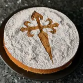 A high-quality, top-down photograph of a traditional Tarta de Santiago, a Spanish almond cake, dusted with powdered sugar in the shape of the Cross of Saint James. The cake is presented on a dark, textured surface, creating a strong visual contrast that highlights the texture and presentation of the dessert.