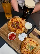A casual, overhead shot of two small pizzas served on a wooden board alongside a pint of Guinness and a bottle of Modelo, likely taken at a pub or casual restaurant.