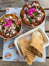 A top-down, overhead shot of three Lebanese street food dishes—a chicken shawarma wrap, a chicken protein box, and a falafel street box—served in cardboard containers on branded paper at an outdoor wooden table.