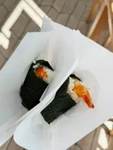 A close-up, top-down shot of two onigiri wrapped in paper, featuring a tempura prawn and a grilled salmon option, set against a wooden background.