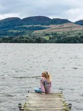 A serene, wide-angle lifestyle shot featuring a person sitting on a wooden jetty, looking out over a calm lake towards rolling hills. The composition uses the jetty as a leading line to draw the viewer's eye into the landscape, creating a peaceful and inviting atmosphere suitable for promoting a getaway or retreat.