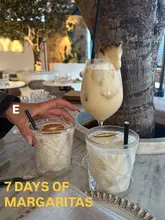 A high-quality, well-composed shot of three cocktails, specifically margaritas, served on a marble table in a stylish venue. The image captures a relaxed, social atmosphere with a tree integrated into the interior design.