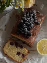 A high-quality, overhead food photography shot of a homemade blackberry and lemon drizzle cake, garnished with fresh blackberries and a dusting of icing sugar. The cake is presented on a cooling rack alongside a fresh lemon slice and a small vase of flowers, creating a rustic and inviting aesthetic.
