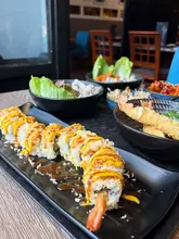 A table spread featuring a variety of Japanese dishes, including a prominent sushi roll in the foreground, rice bowls, and tempura, showcasing a restaurant's menu.